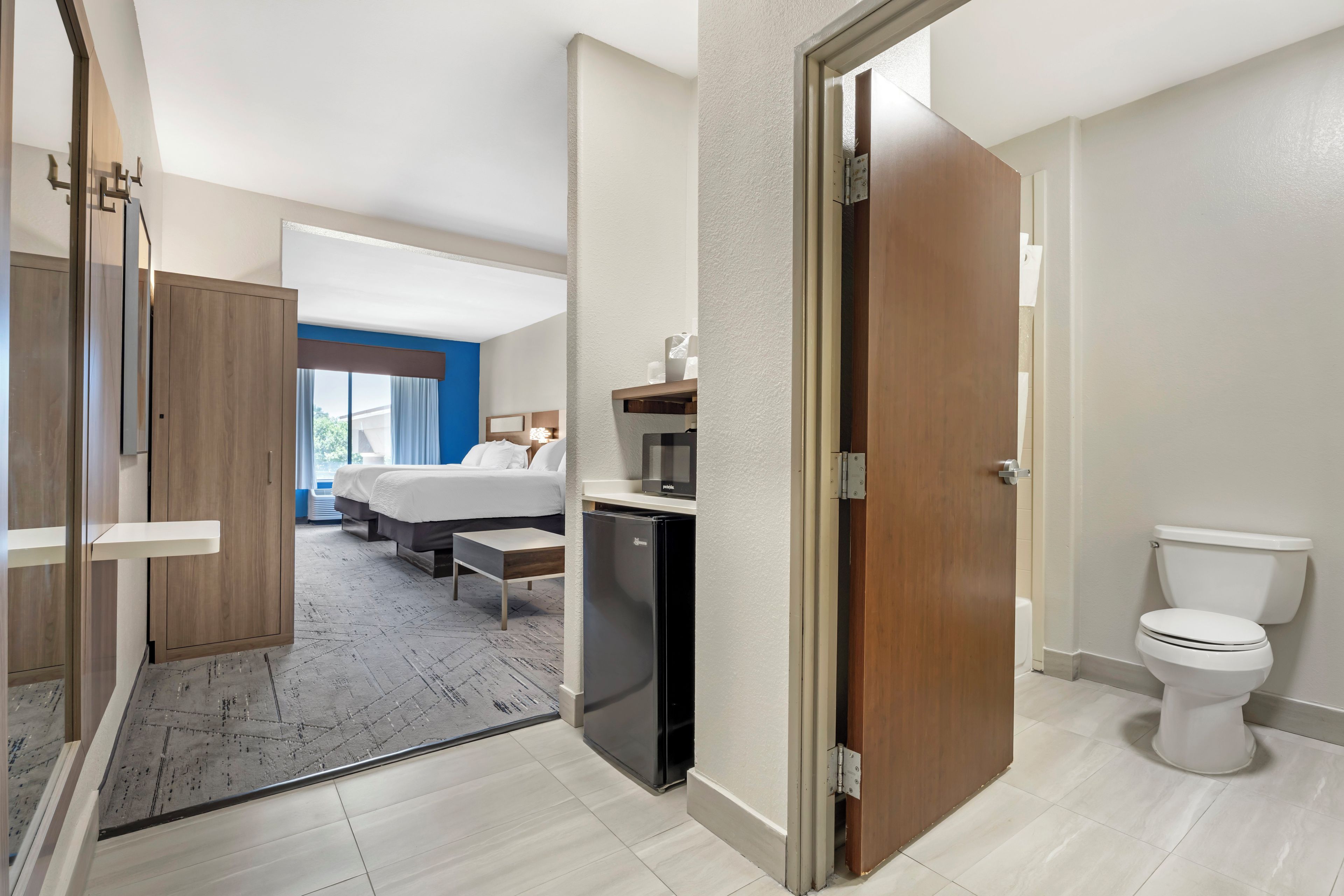 A hotel room with a bed, a small refrigerator, a bathroom with a toilet, and a view of the outside.