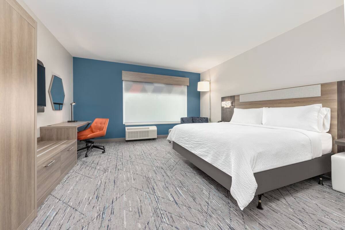 A modern hotel room with a large bed, a desk with a chair, a window with blinds, and a floor lamp.