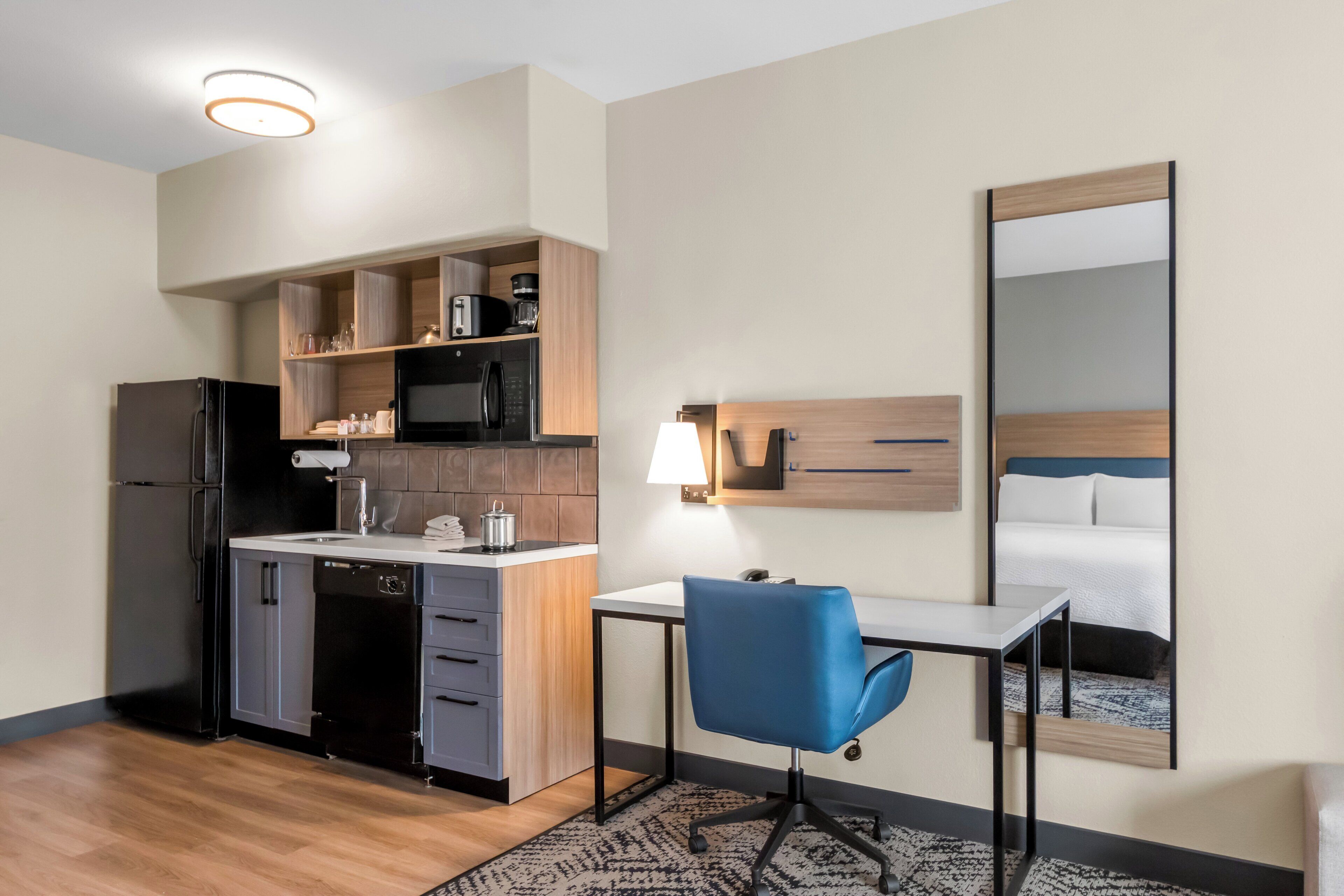 A compact hotel room with a kitchenette, a desk, a chair, a bed, and a large mirror.