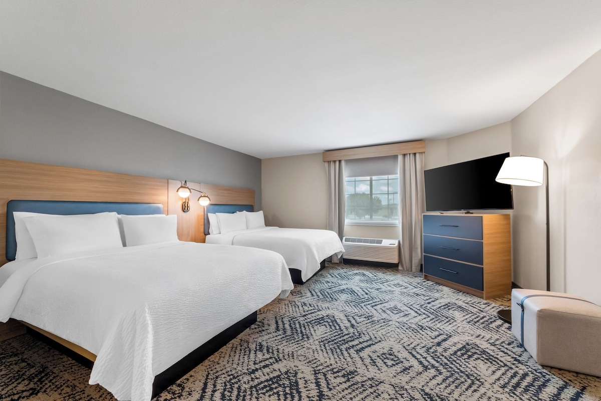 A hotel room with two beds, a flat-screen TV, a desk with drawers, a lamp, and a large window with curtains.