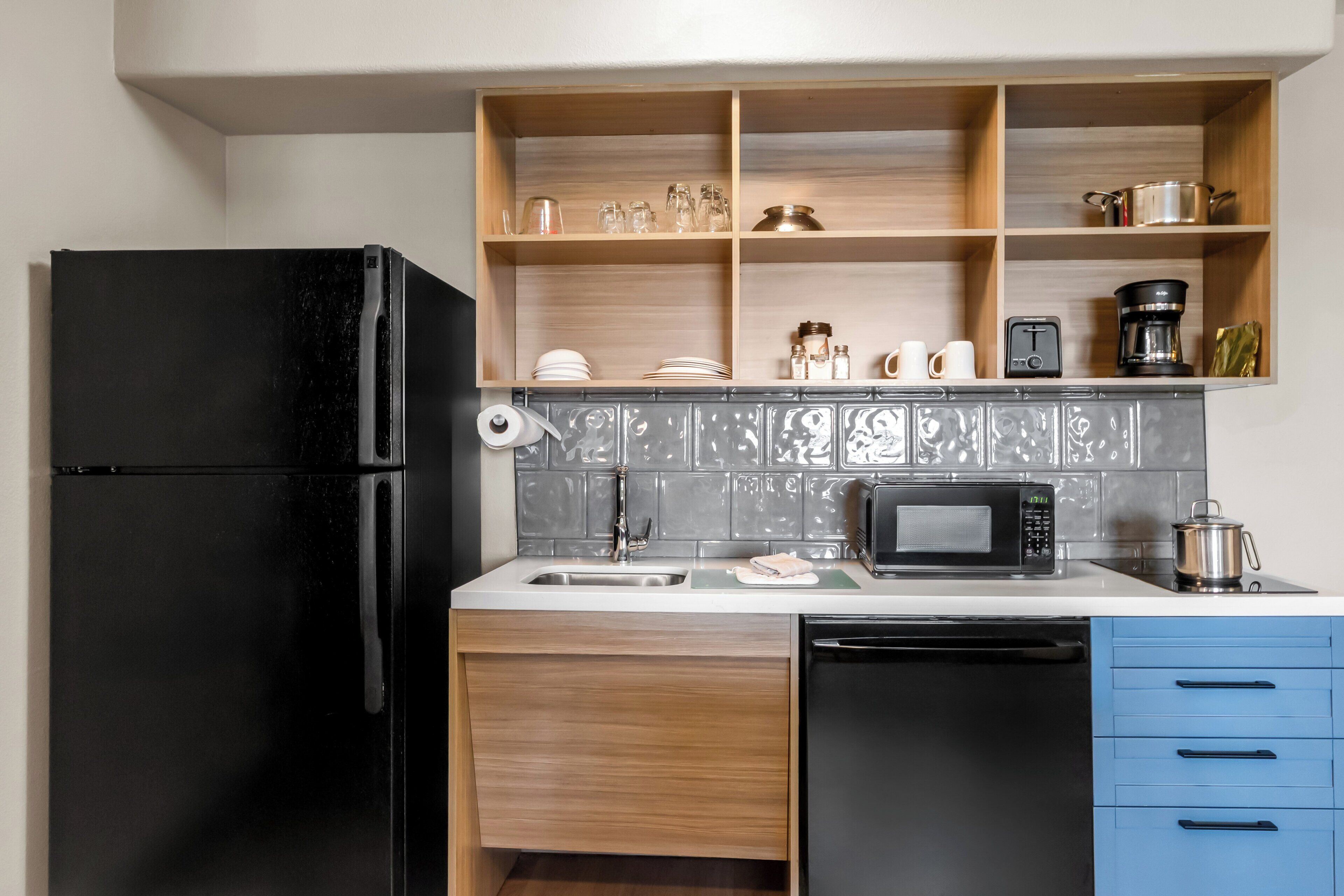 A compact kitchen with a black refrigerator, a microwave, a sink, and a dish rack. There are wooden shelves above the counter holding various kitchen items.