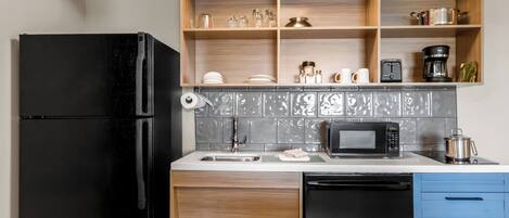 A compact kitchen with a black refrigerator, a microwave, a sink, and a dish rack. There are wooden shelves above the counter holding various kitchen items.
