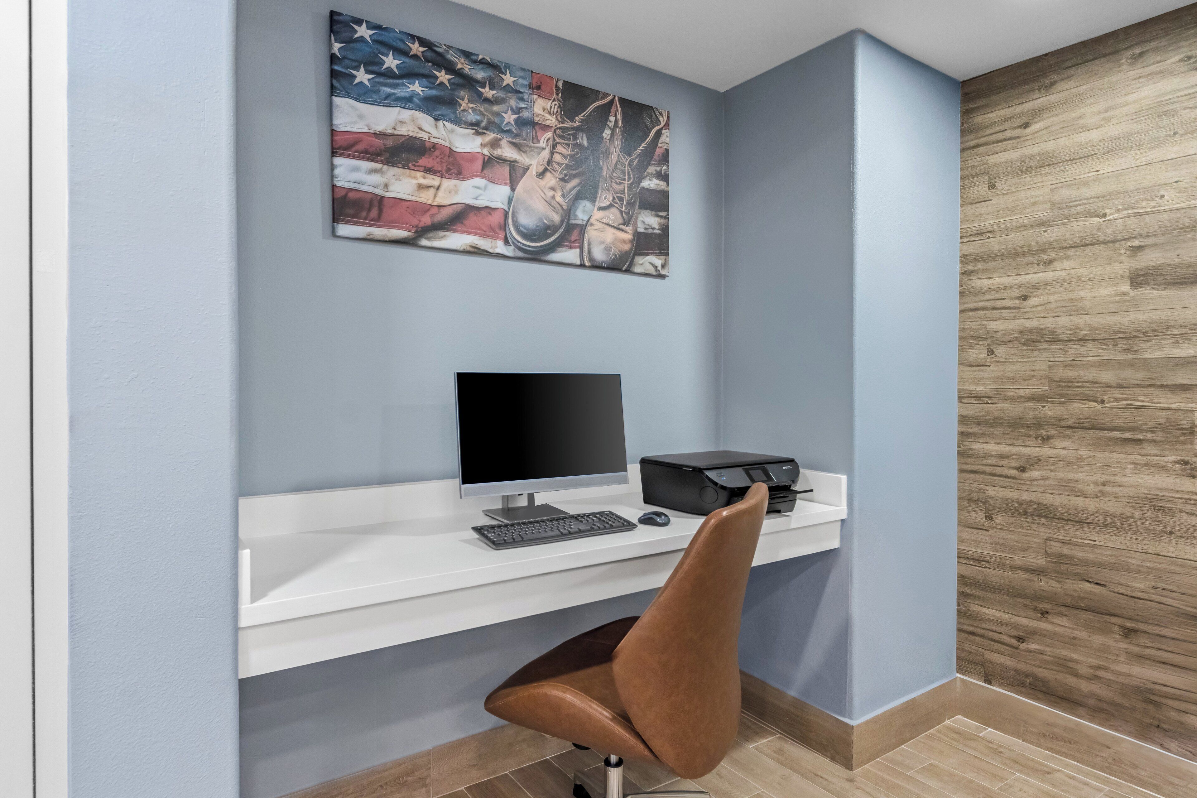 A modern office desk with a computer, keyboard, and mouse, a brown leather chair, and a wall-mounted American flag painting.