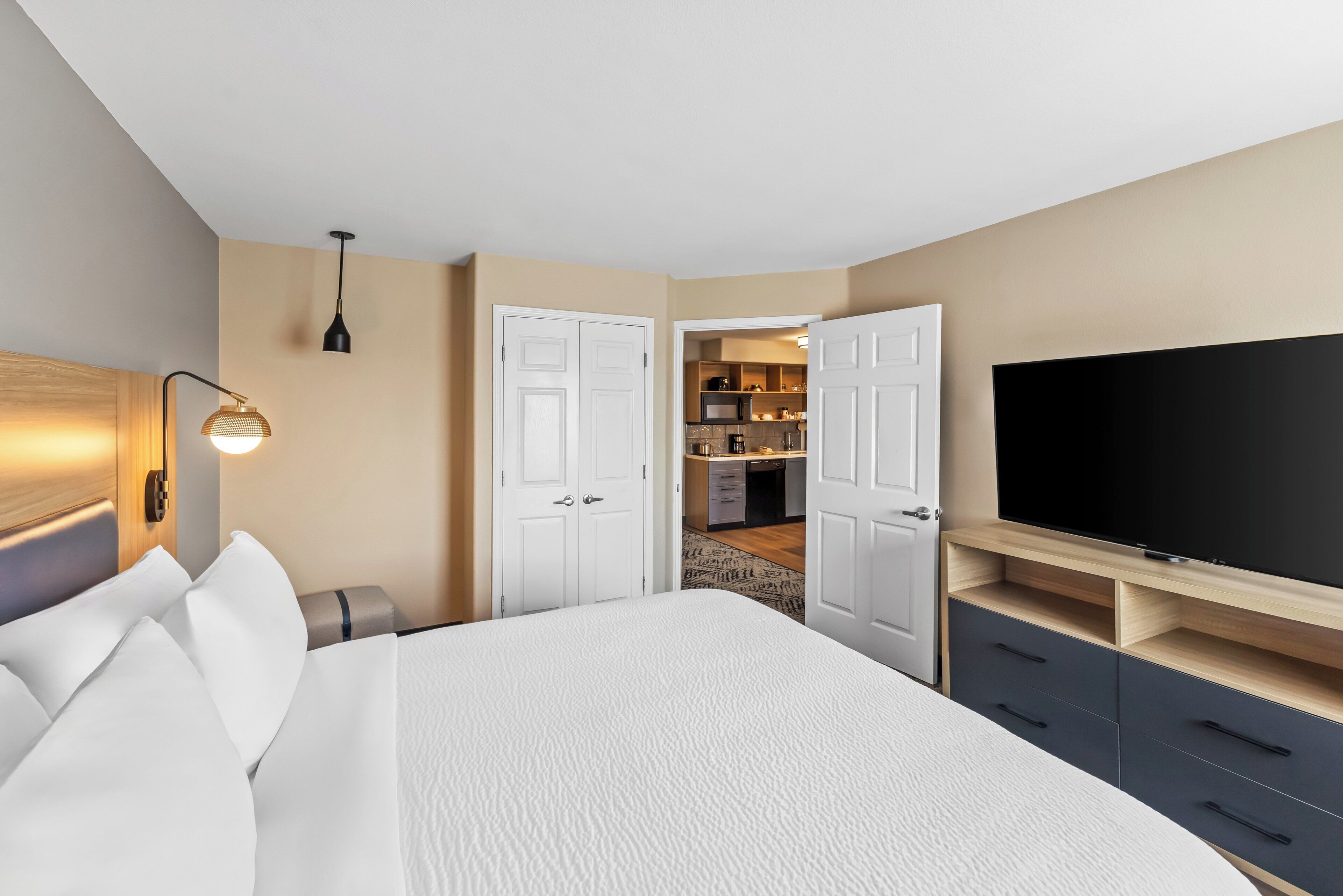 A hotel room with a bed, a TV, a desk, and a kitchenette in the background.