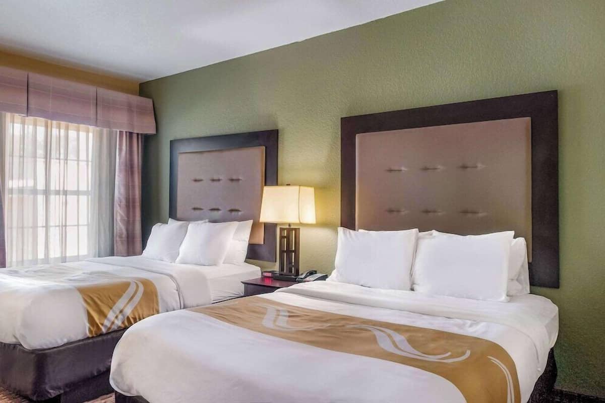 A hotel room with two beds, a nightstand, a lamp, and a window with curtains.