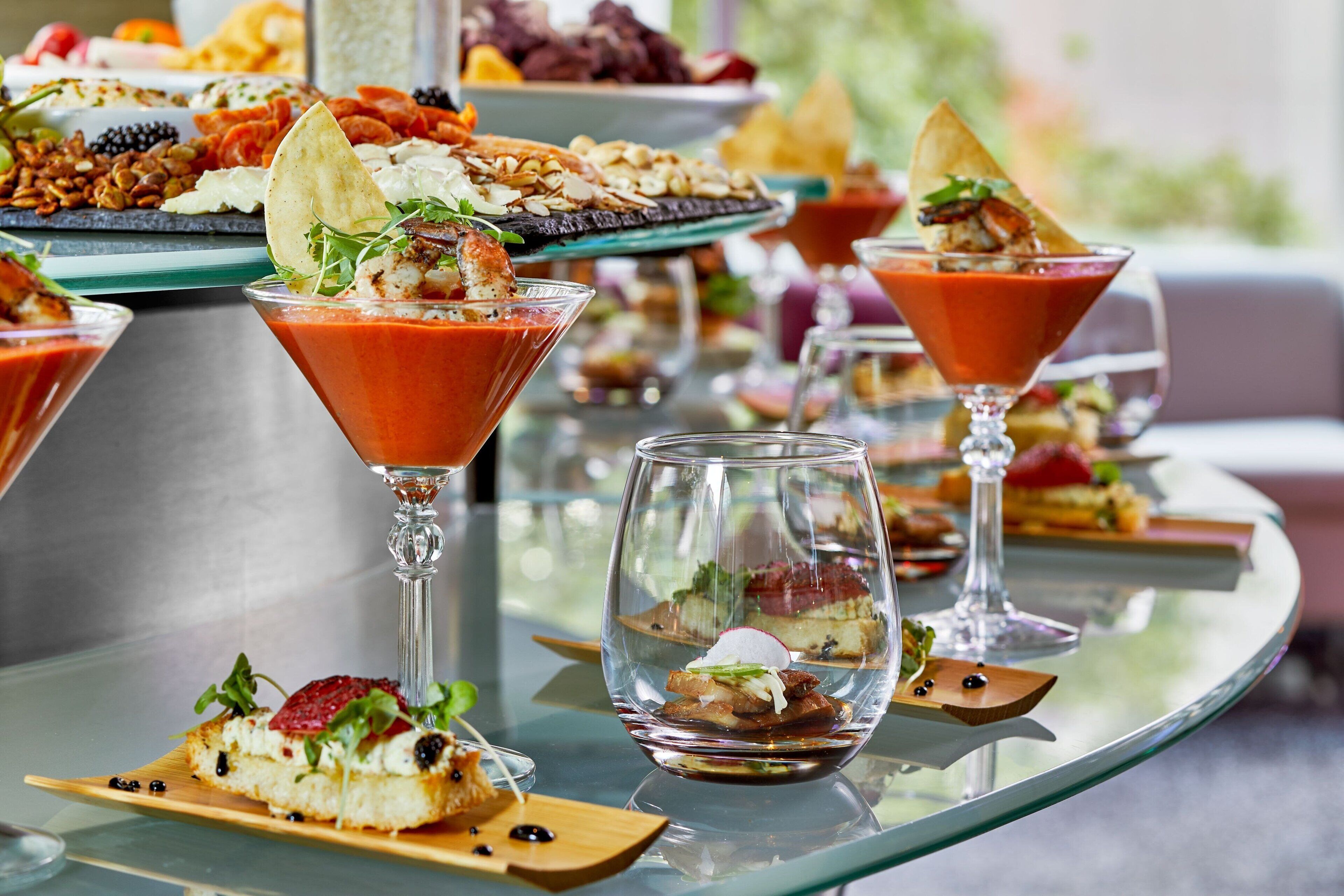 Une table recouverte d’une vitre, sur laquelle sont disposés divers aliments et boissons, notamment un verre à martini contenant une boisson rouge et un petit bol rempli d’un dessert.