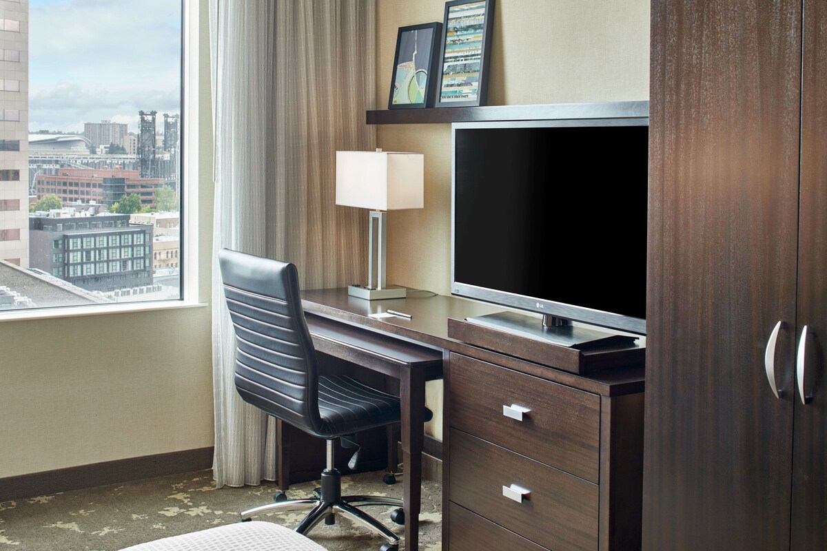 A hotel room with a desk, chair, TV, and a view of the cityscape through the window.
