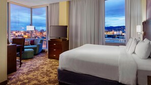 A hotel room with a large bed, a TV, a sofa, and a view of a cityscape at dusk. - Courtyard by Marriott Portland City Center (Portland)