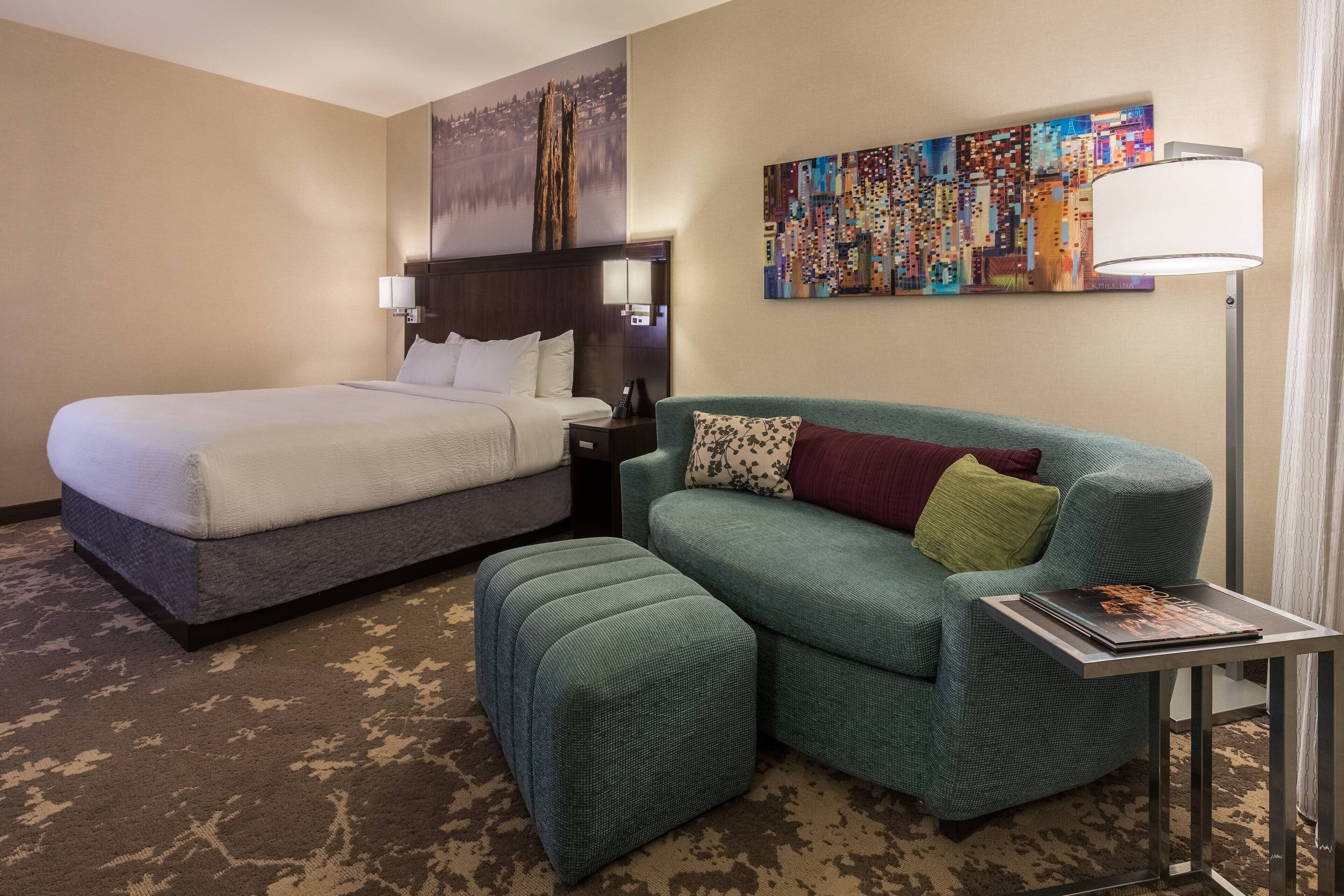 A hotel room with a bed, a sofa, a small table with a lamp, and a painting on the wall.