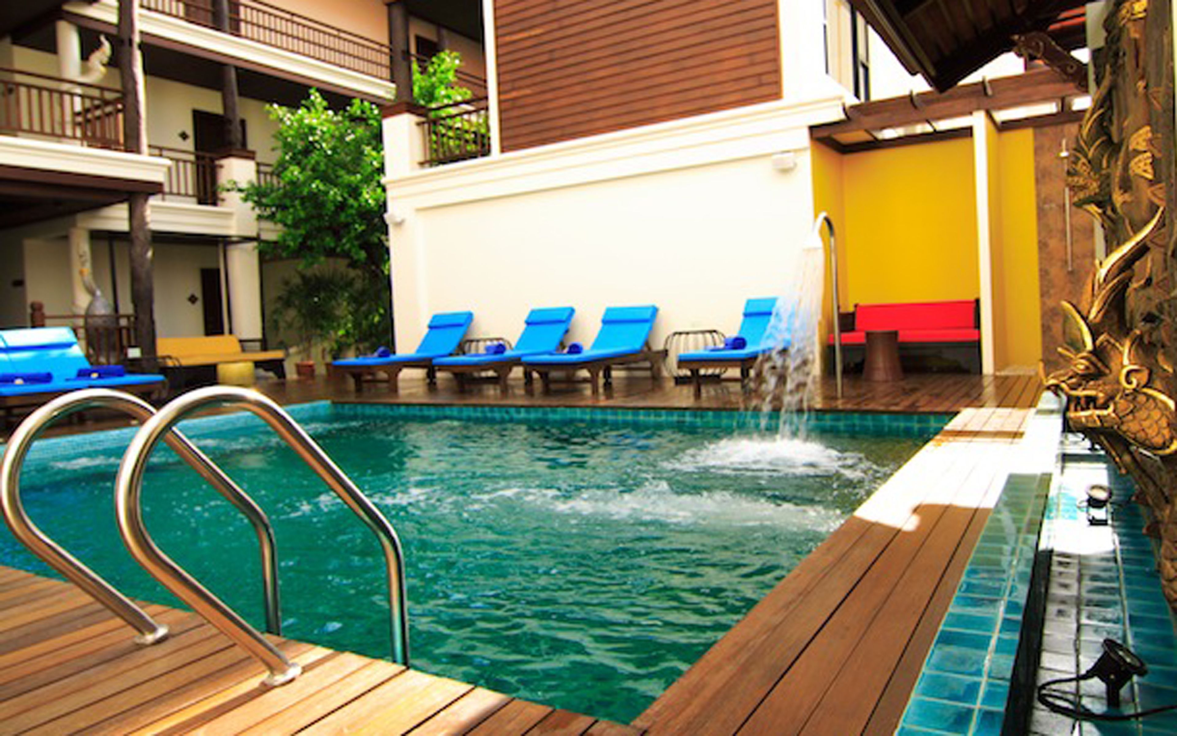 A pool area with a wooden deck, blue lounge chairs, and a water feature.