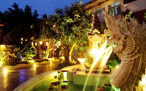 Un jardín nocturno con fuente ornamental, estatuas iluminadas y un edificio tradicional al fondo.