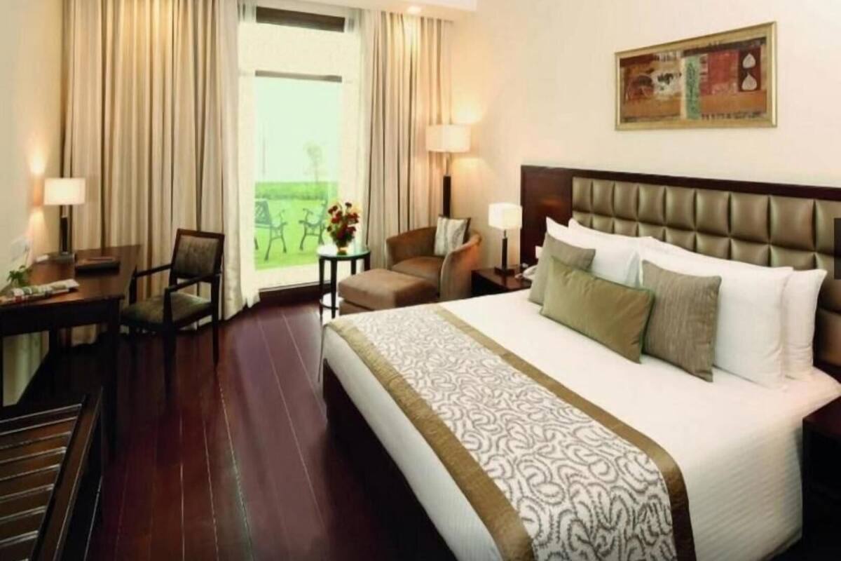 A hotel room with a large bed, a desk, a chair, a small table with a lamp, and a view of the outdoors.