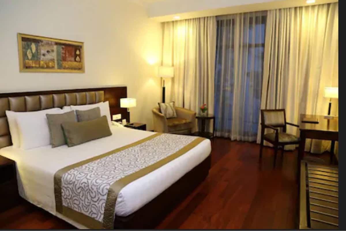 A hotel room with a large bed, a headboard, a desk, a chair, a lamp, and a painting on the wall.