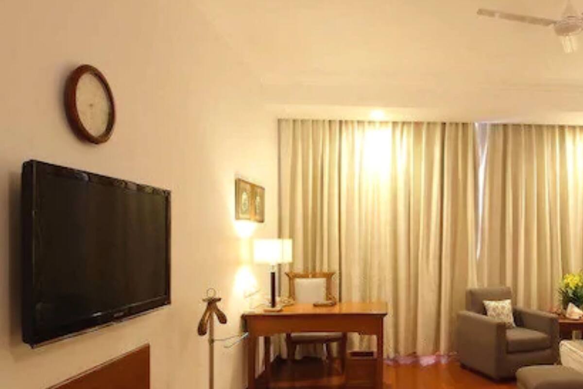 A hotel room with a flat-screen TV, a desk with a lamp, a chair, and a large window with curtains.