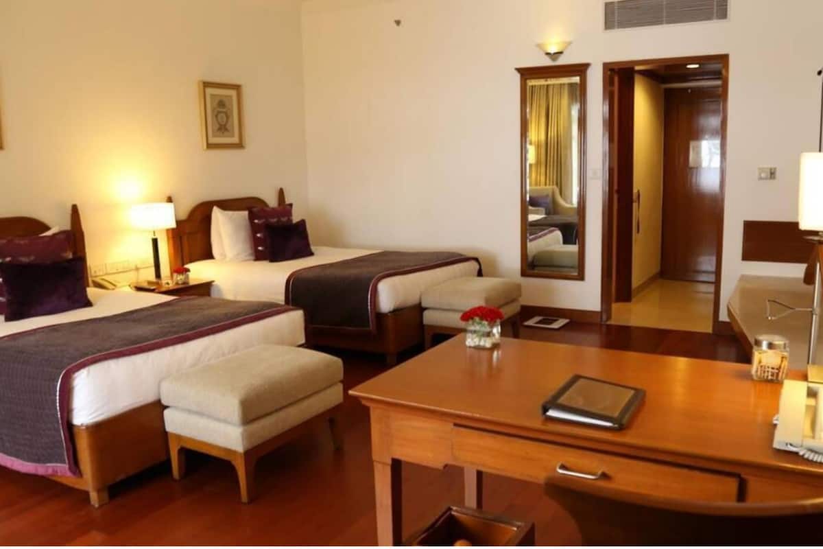 A hotel room with two beds, a desk with a chair, a small table with a vase of flowers, and a mirror on the wall.