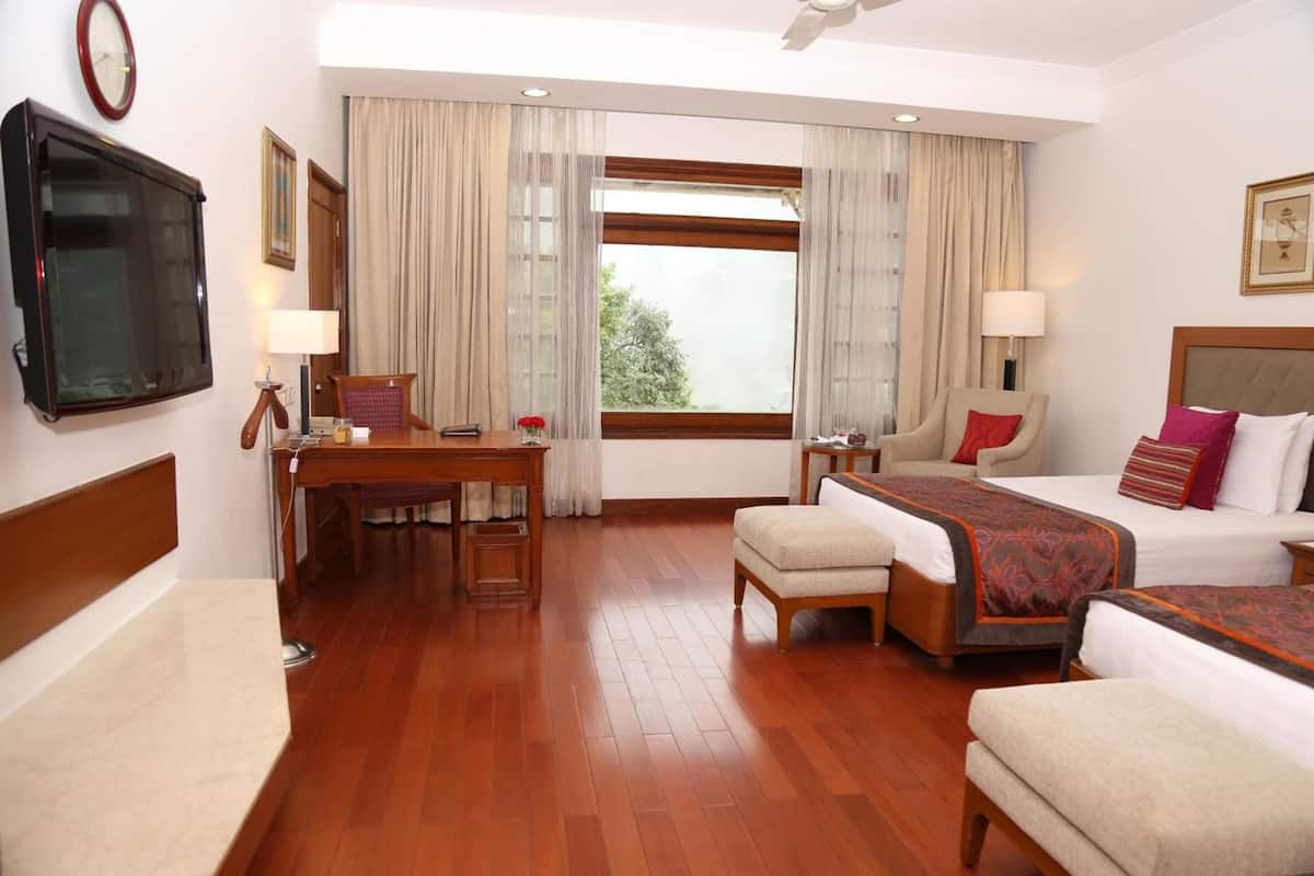A hotel room with a wooden floor, two beds, a desk, a chair, a TV, and a window with curtains.