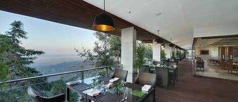A dining area with glass tables and wicker chairs, overlooking a mountainous landscape.