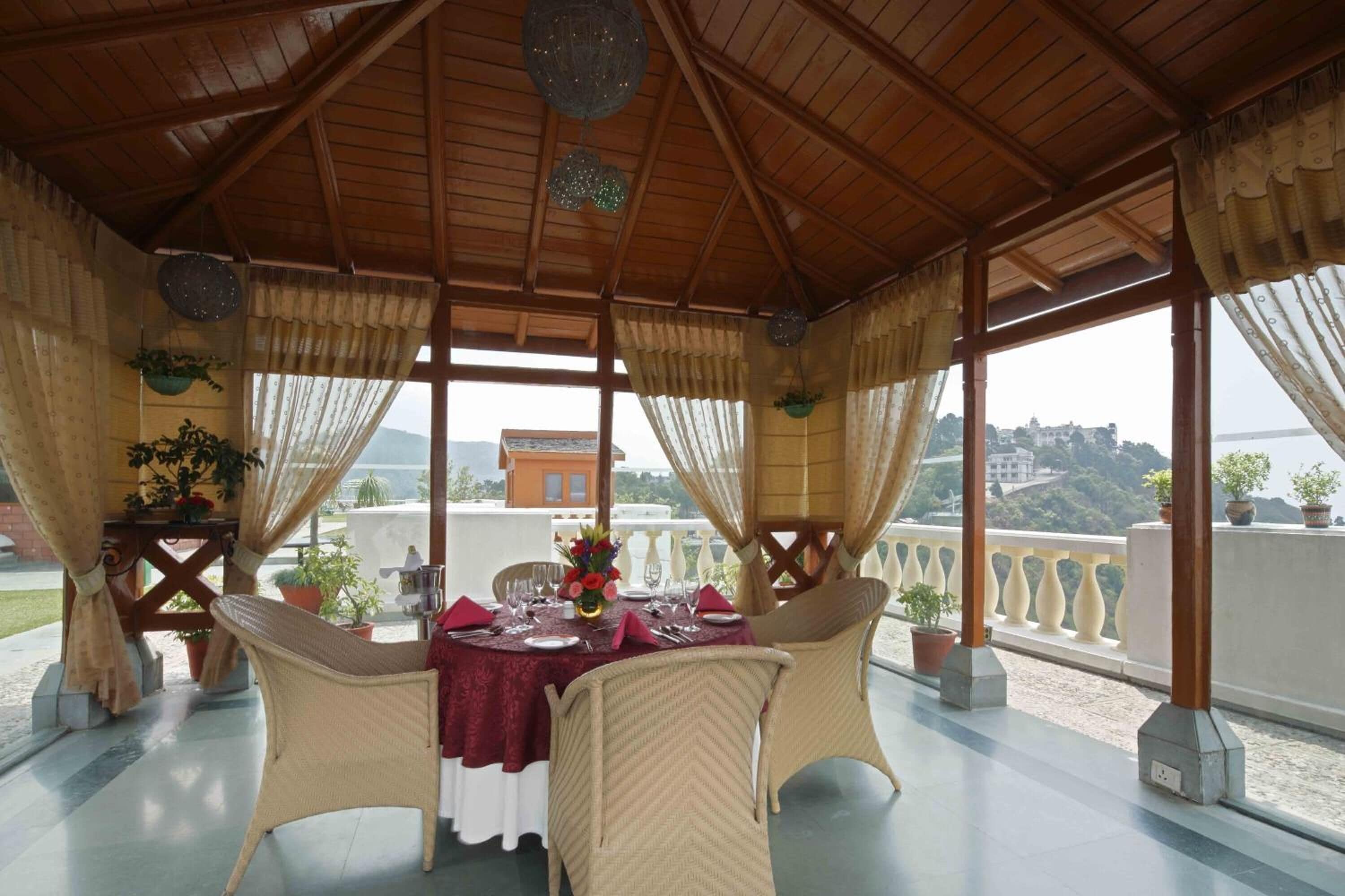 Uma área de refeições ao ar livre com uma mesa posta para dois, rodeada por cadeiras de vime, sob um telhado de madeira com vista para a cidade.