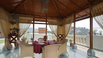 Un área de comedor al aire libre con una mesa preparada para dos personas, rodeada de sillas de mimbre, bajo un techo de madera con vista a la ciudad.