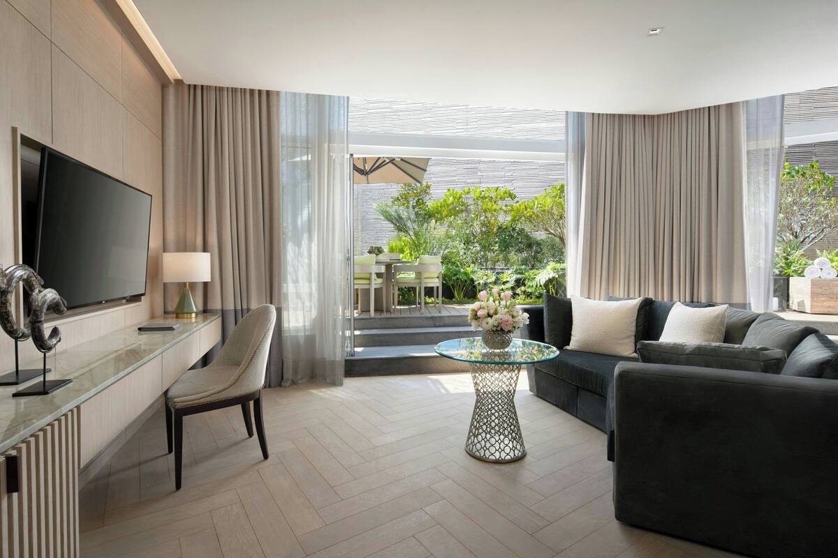 A modern hotel room with a flat-screen TV, a glass coffee table with a floral centerpiece, a sofa, and a dining area visible through large windows.