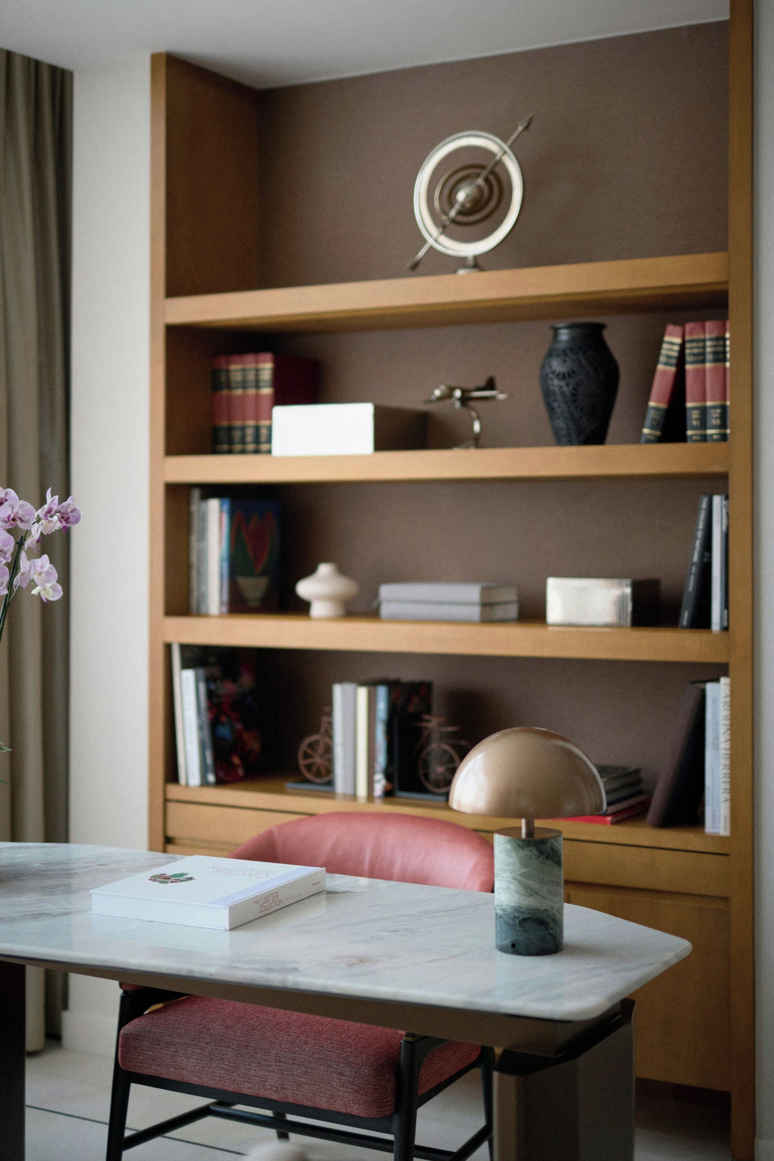 Una habitación con una mesa de mármol, una silla tapizada en rojo, una estantería con libros y adornos, y un jarrón con flores.