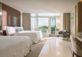 A hotel room with two beds, a sitting area with a chair, a small table, and a balcony with a view.