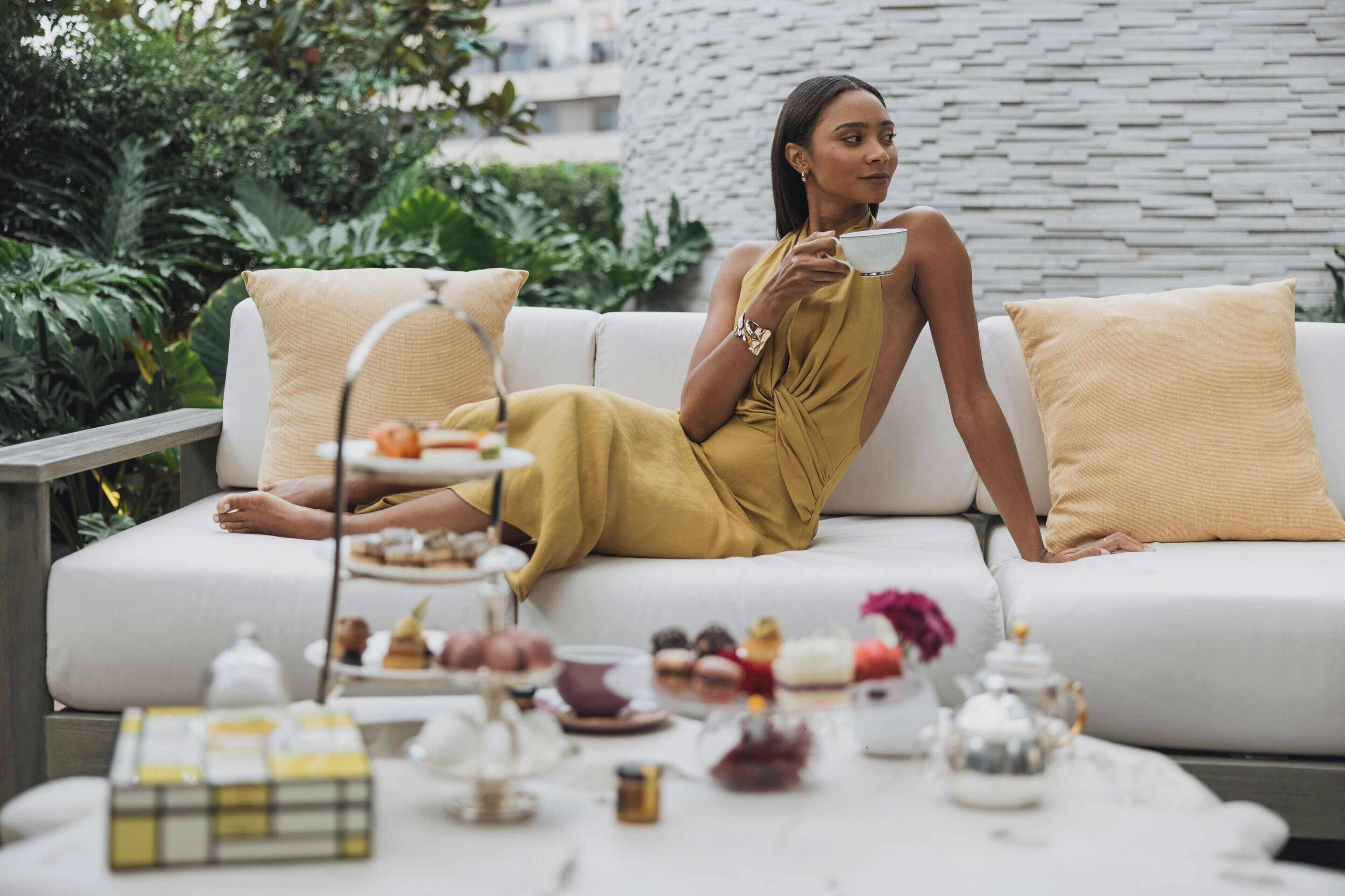 A woman sitting on a white sofa with a tray of pastries and tea set in front of her.