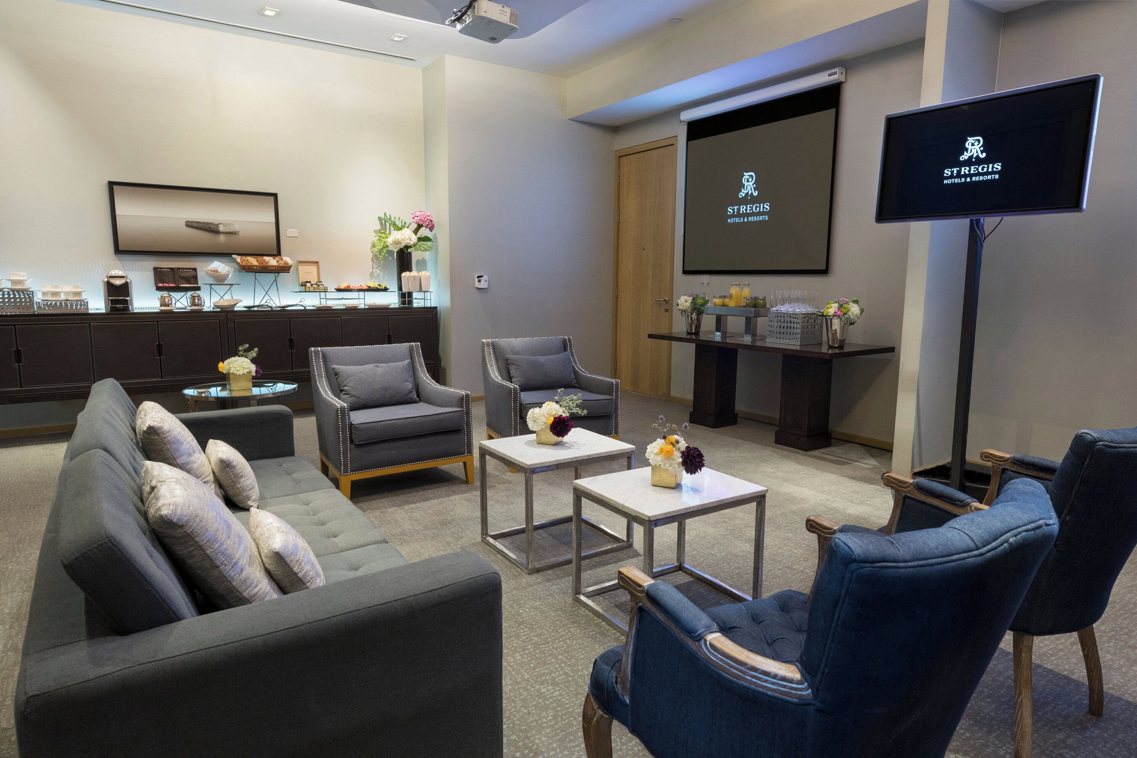 Een hotel lobby met een grijze bank, fauteuils, een bijzettafeltje en een televisiescherm waarop de naam van het hotel staat.
