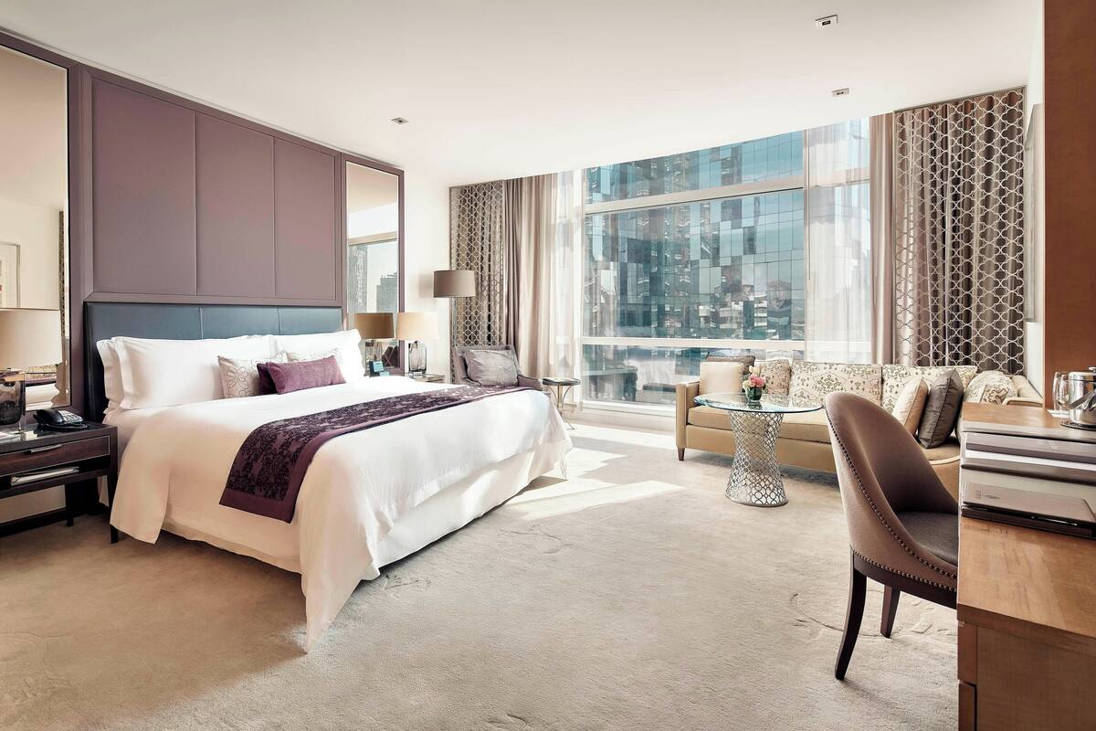 A modern hotel room with a large bed, a sofa, a desk, and a view of the city through large windows.