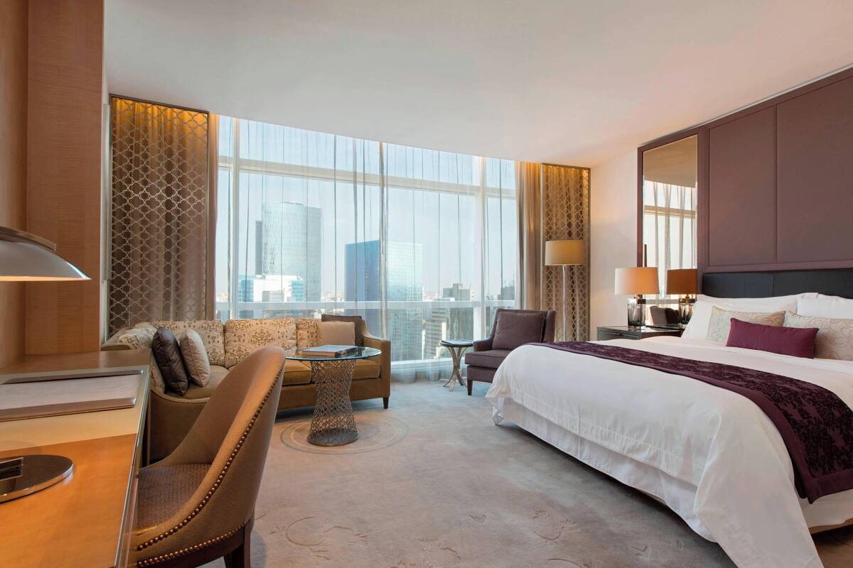 A hotel room with a large bed, a sitting area with a sofa and chairs, a small round table, and a large window offering a city view.