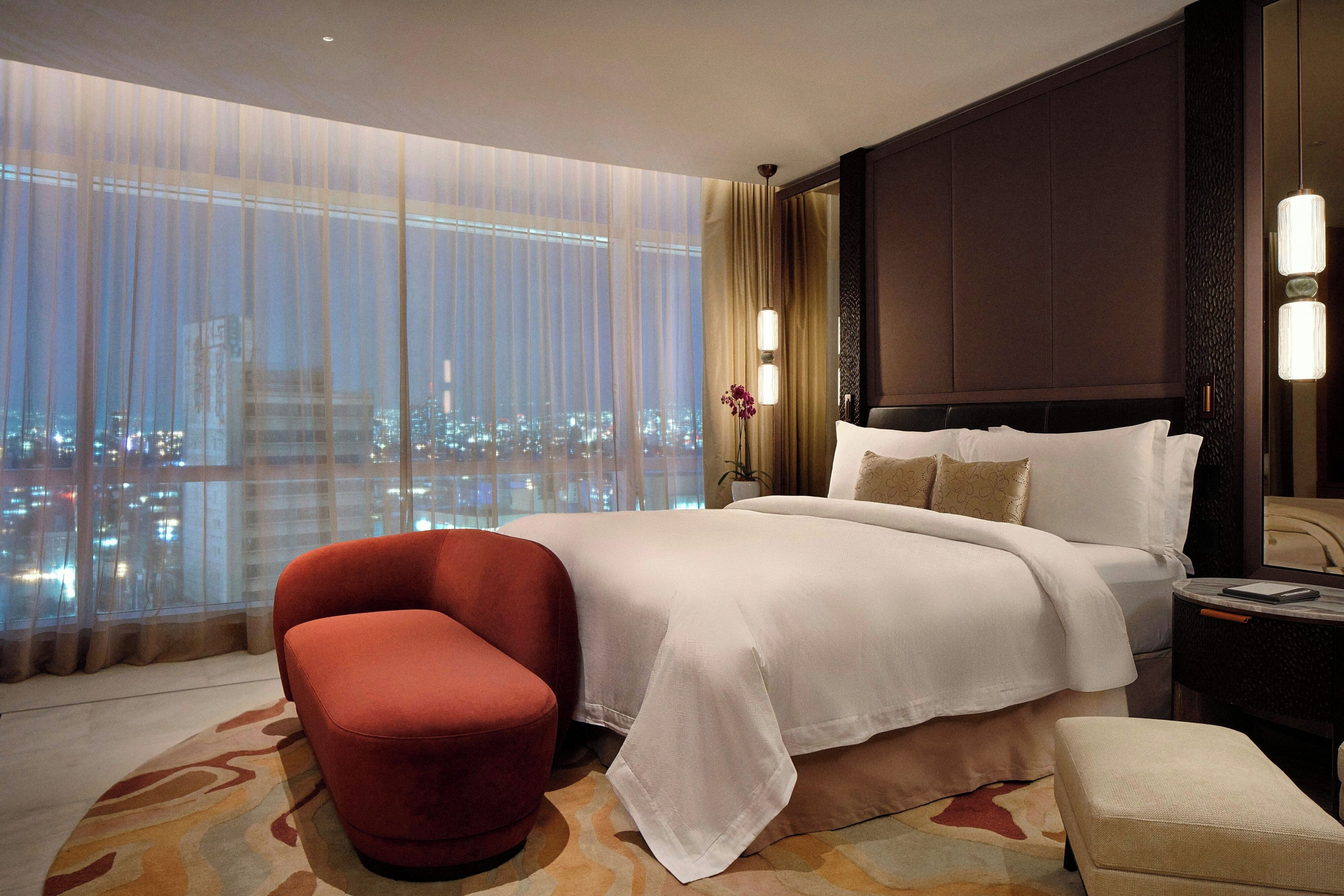 A hotel room with a large bed, a red armchair, a nightstand, and a view of a cityscape through floor-to-ceiling windows.