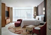 A hotel room with a large bed, a TV, a desk, a chair, a small table, and a view of the city through large windows.