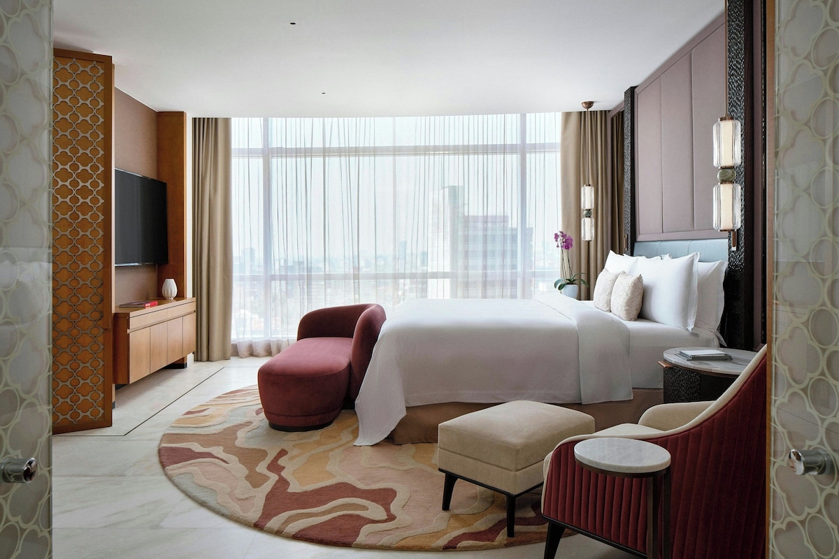 Habitación de hotel con una cama grande, televisión, escritorio, silla, mesita y vista a la ciudad a través de amplios ventanales.