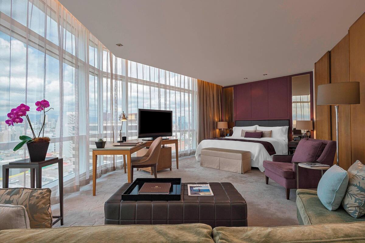 A modern hotel room with a large window, a bed, a desk with a TV, a sofa, and a coffee table.