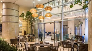 A modern dining area with multiple tables and chairs, large hanging lamps, and a glass partition separating the dining space from the rest of the building.