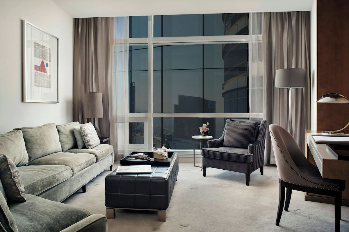 A modern living room with a sofa, armchair, ottoman, and a small table with a lamp.
