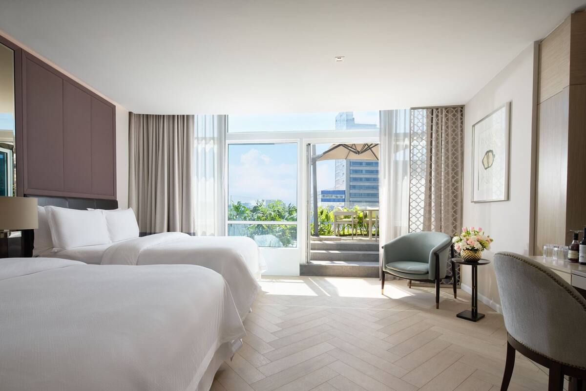 A hotel room with two beds, a desk, a chair, and a balcony with a view of the city.