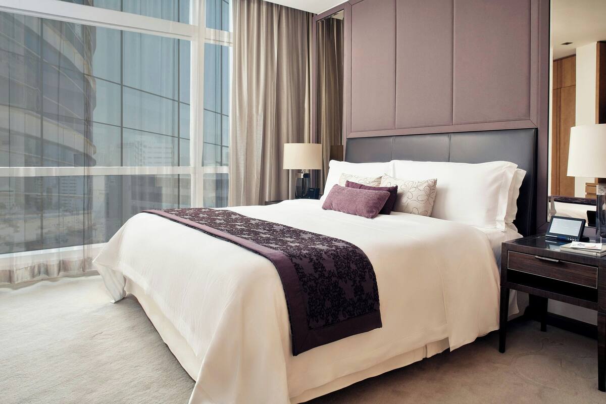 A hotel room with a large bed, a nightstand, a lamp, and a view of a building through the window.