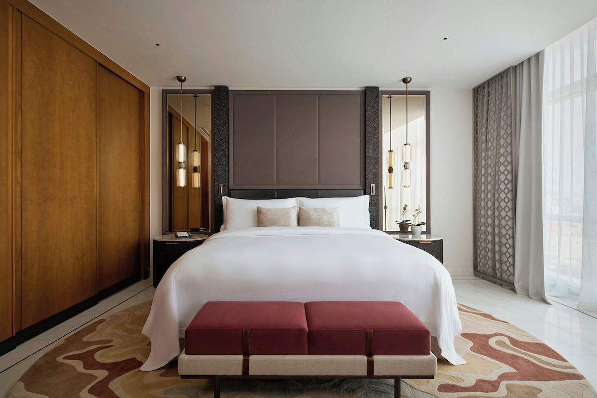 A modern hotel room with a large bed, a bench, a nightstand, and a large mirror.