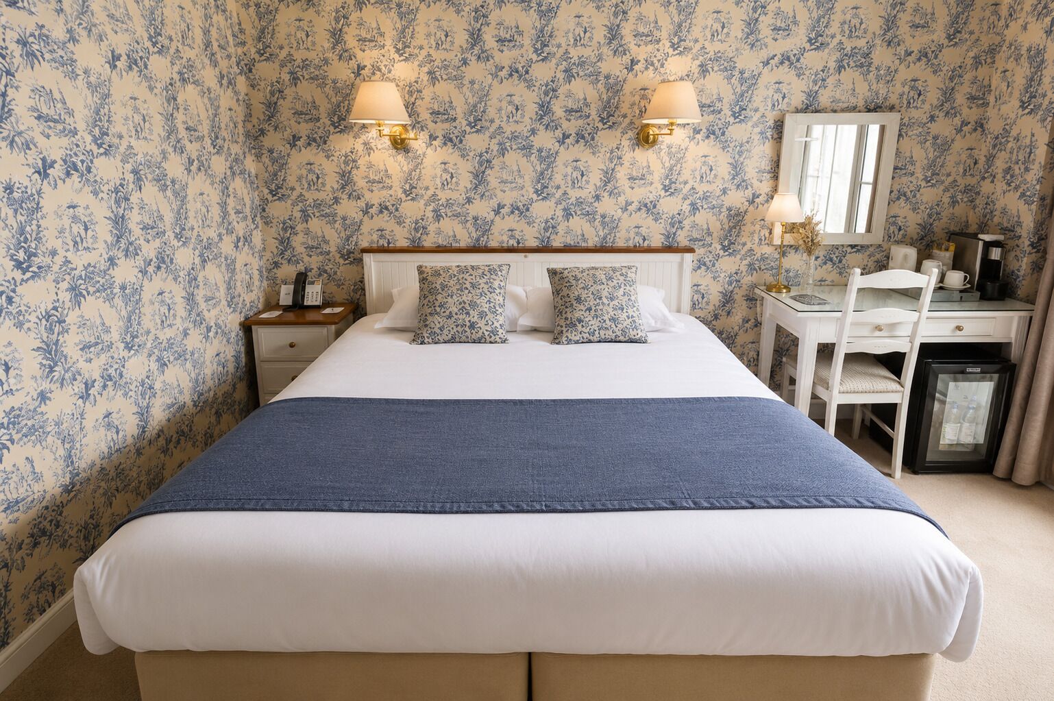 A hotel room with a bed, bedside tables, a desk, a chair, a mirror, and a refrigerator.