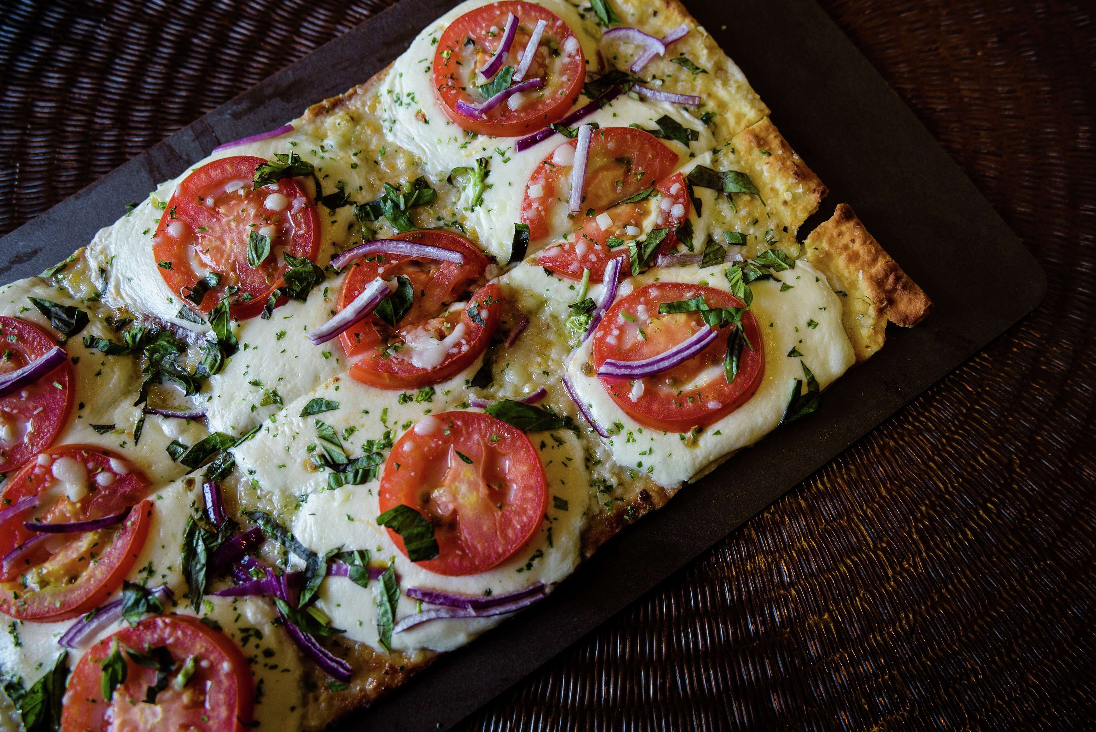 En rektangulær pizza med tomatsskiver, rødløg og krydderurter på en mørk skiferplade.