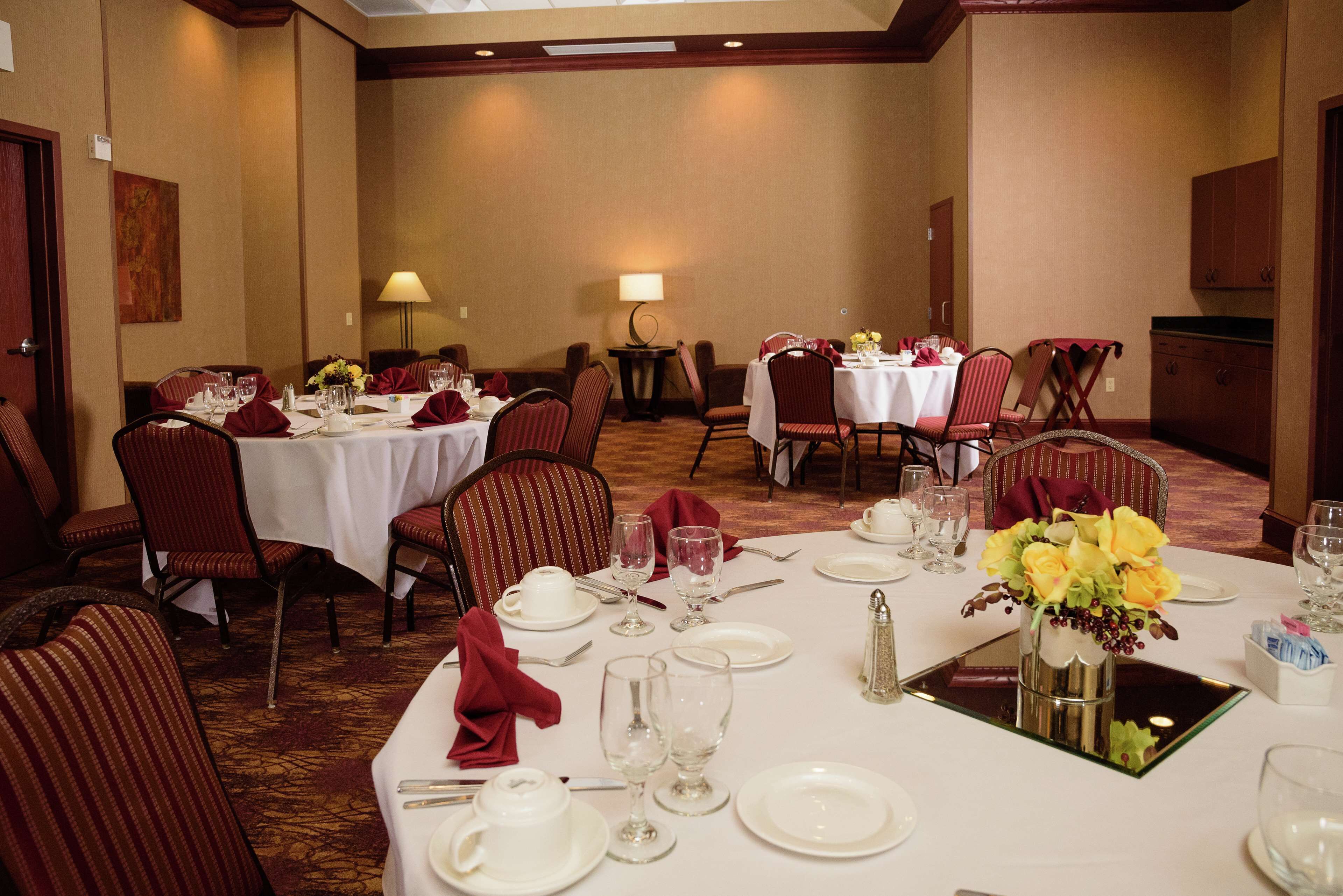 Comedor con mesas dispuestas para cuatro personas, cada una con mantel blanco, servilletas rojas y centros de mesa florales.