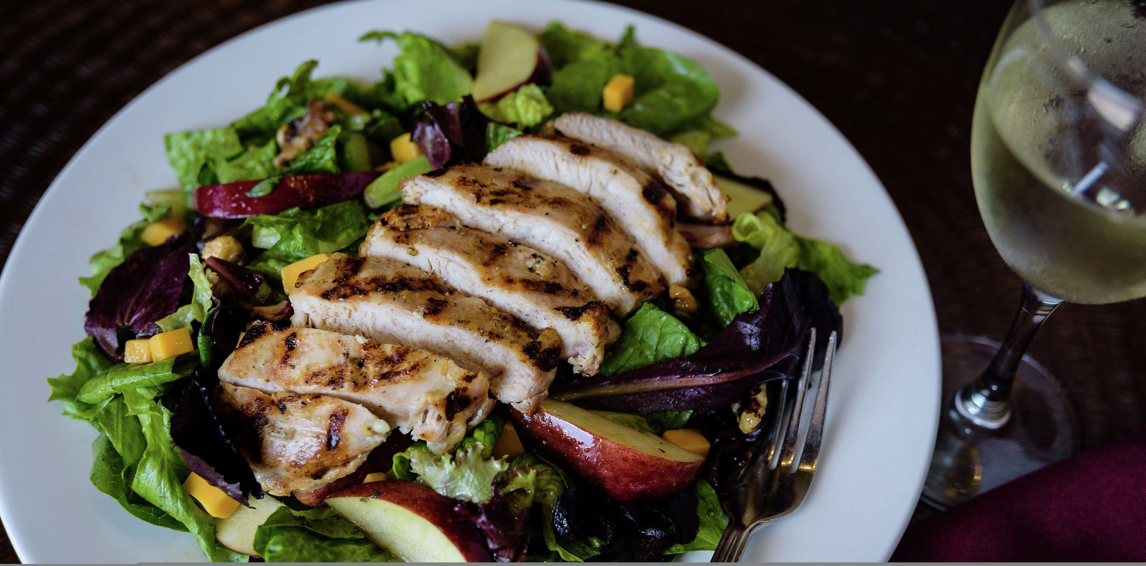 A plate of grilled chicken and salad with a glass of white wine on the side.