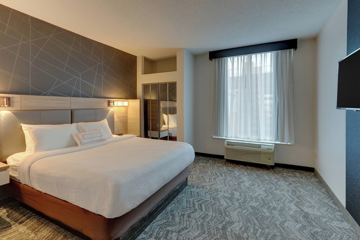 A hotel room with a large bed, a nightstand, a mirror, a window with curtains, and a flat-screen TV mounted on the wall.