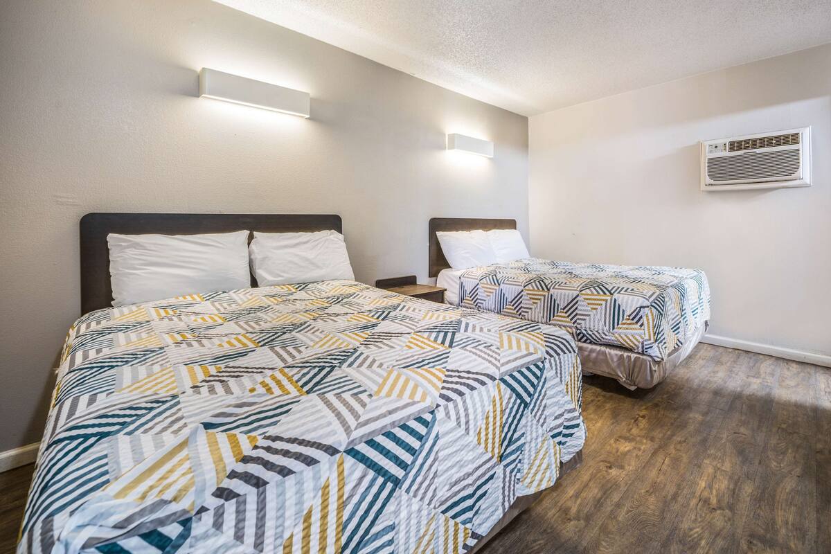 Habitación de hotel con dos camas, cada una con una colcha estampada, una mesita de noche, aire acondicionado montado en la pared y suelo de madera.