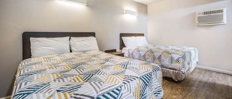 A hotel room with two beds, each with a patterned bedspread, a nightstand, a wall-mounted air conditioner, and a wooden floor.