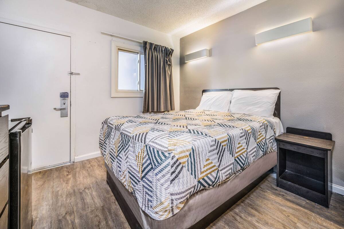 Un dormitorio con cama, mesita de noche, ventana con cortinas y puerta.