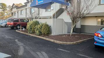 Una entrada de hotel con un letrero de '6 welcome', un estacionamiento con varios coches y una pequeña zona ajardinada con arbustos y un árbol.