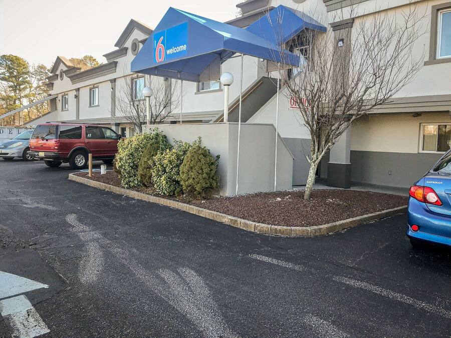 Una entrada de hotel con un letrero de '6 welcome', un estacionamiento con varios coches y una pequeña zona ajardinada con arbustos y un árbol.