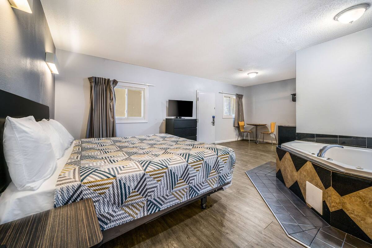Habitación de hotel con cama, televisor, baño con bañera y un pequeño rincón de descanso.